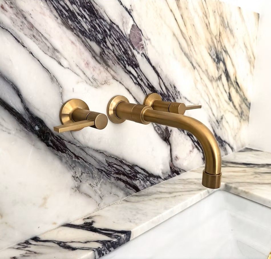 Gold wall-mounted faucet against white and gray marble backsplash, above a white sink or tub edge