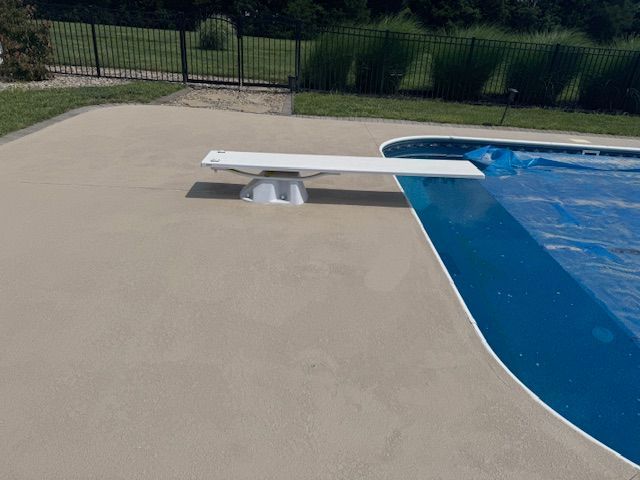 A tan concrete pool deck with a white diving board extending over a blue-covered pool.