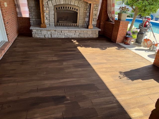 Wood-look concrete patio with a stone fireplace, adjacent to a pool and brick wall.