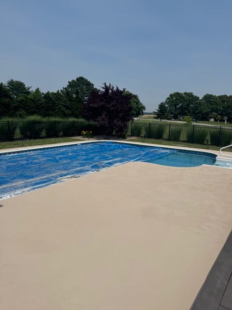 Pool with tan deck partially covered by a blue pool cover, surrounded by greenery and a blue sky.