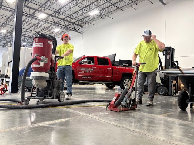 Two workers grinding a concrete floor in a warehouse, using machinery. Red truck in background.