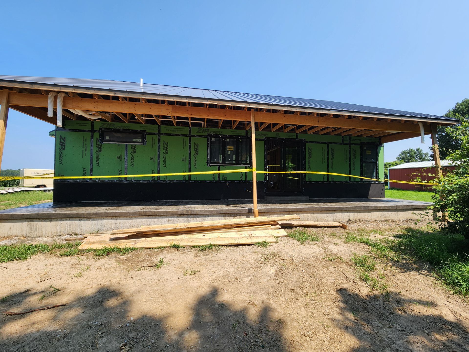 Construction site of a single-story building; wooden frame, green sheathing, dark siding, gray roof, yellow caution tape.