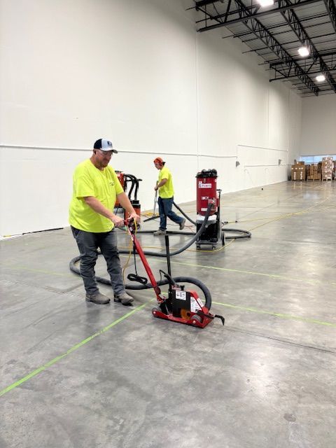 Two workers in a warehouse using floor grinders and vacuums. Green tape lines the concrete floor.