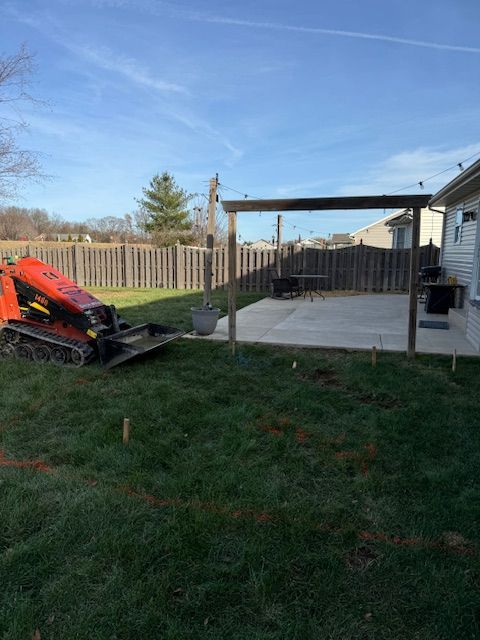 Small construction equipment and a new patio in a grassy backyard, framed by a wooden pergola; blue sky.