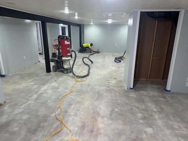 Basement renovation with a worker using an industrial vacuum. Concrete floor and gray walls.