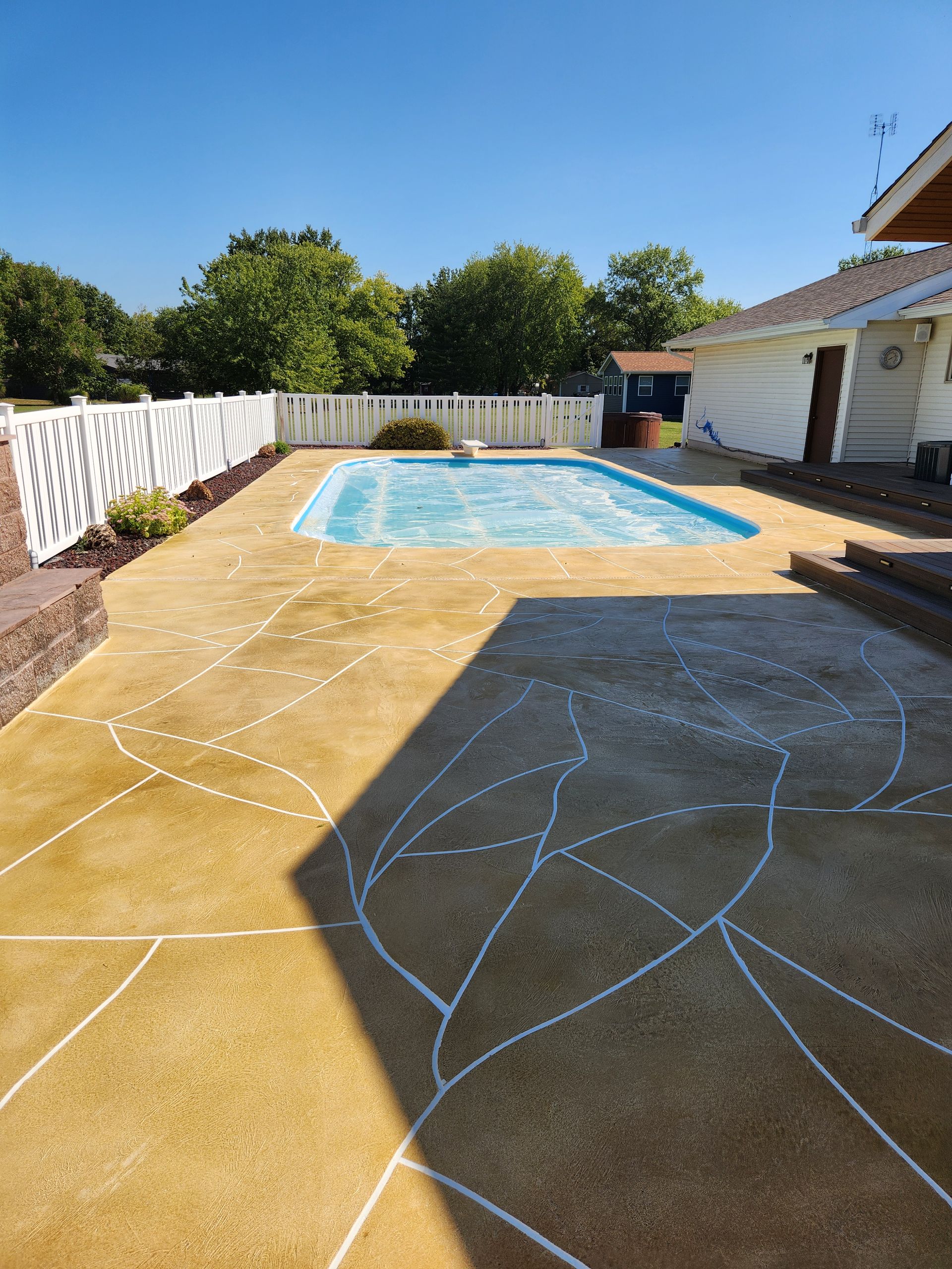 Backyard with pool, tan patio, white lines, white fence, trees, and house. Blue sky.