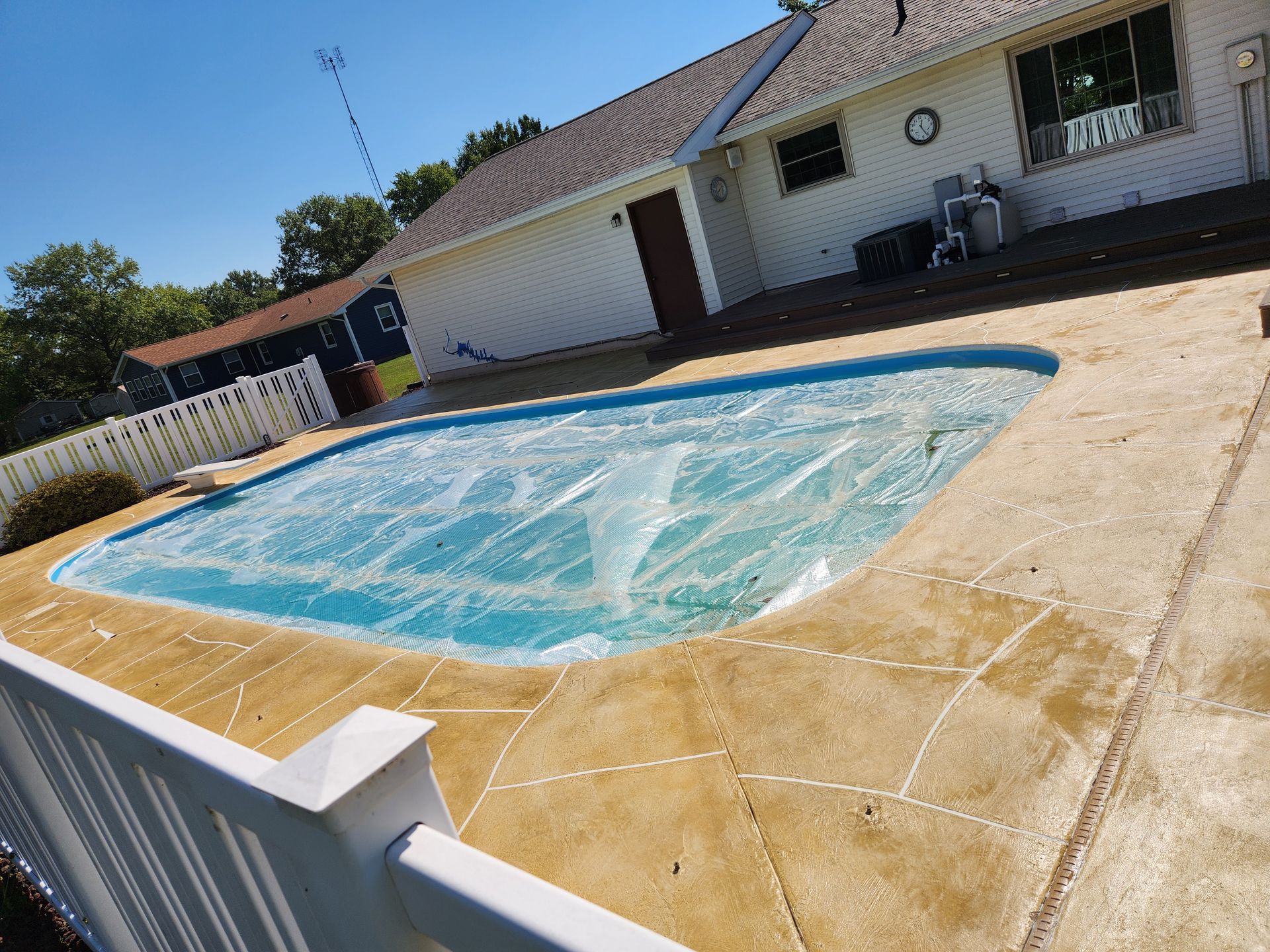 Pool covered with a blue tarp, surrounded by a tan concrete patio and a white fence, under a blue sky.