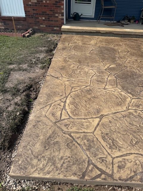Stamped concrete patio with stone pattern, bordered by grass and a brick house.
