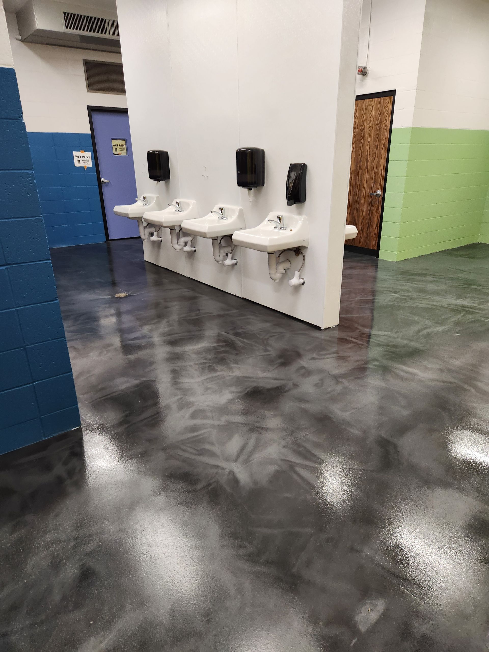 Bathroom with three sinks, black soap dispensers, and shiny gray floor. Walls are white, blue, and green.