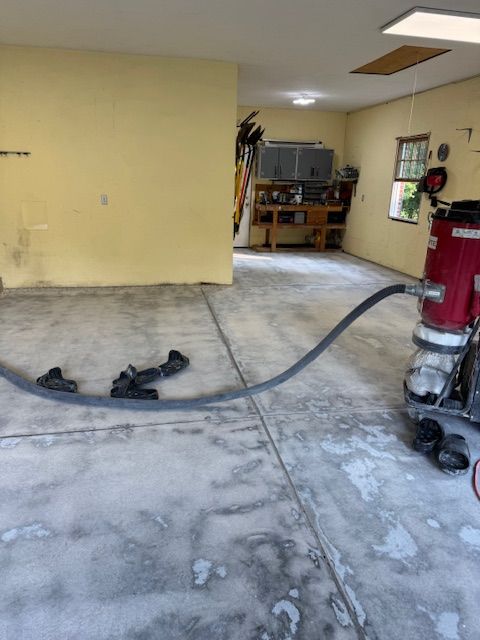 Garage floor being prepared, with equipment connected by a hose.  Gray concrete, yellow walls, and a tool bench in the background.