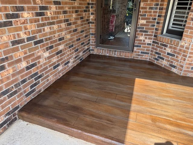 Brick porch with a wood-grain stained concrete floor next to a brick wall and door.