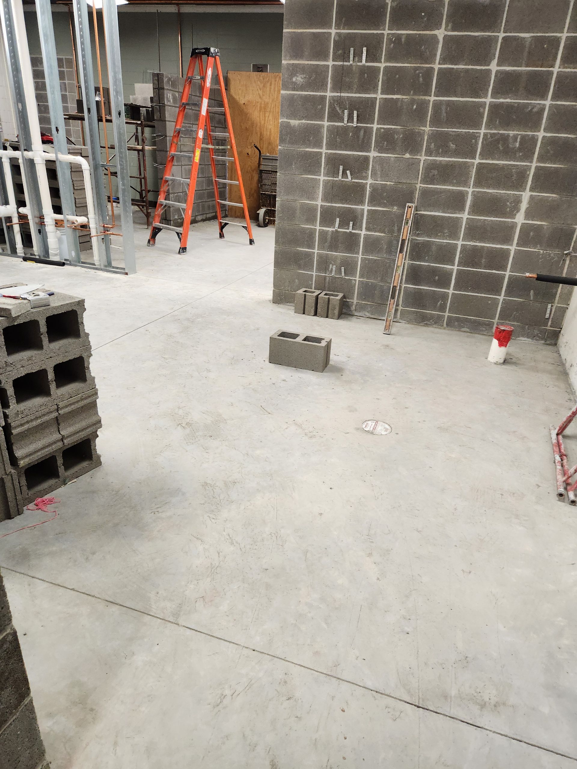 Concrete block construction site, with partially built wall, ladder, and supplies on concrete floor.