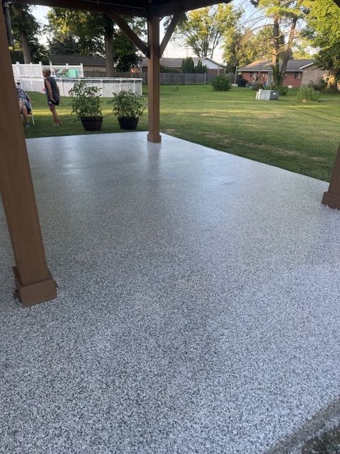 Gray speckled concrete patio under a wooden pergola, overlooking a grassy yard with houses in the background.