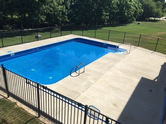 A rectangular blue swimming pool with a black fence, surrounded by concrete and grass.