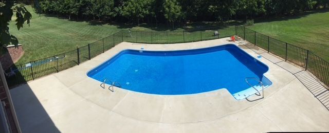 Overhead view of a blue swimming pool surrounded by a concrete patio and a black fence. Green grass and trees are in the background.