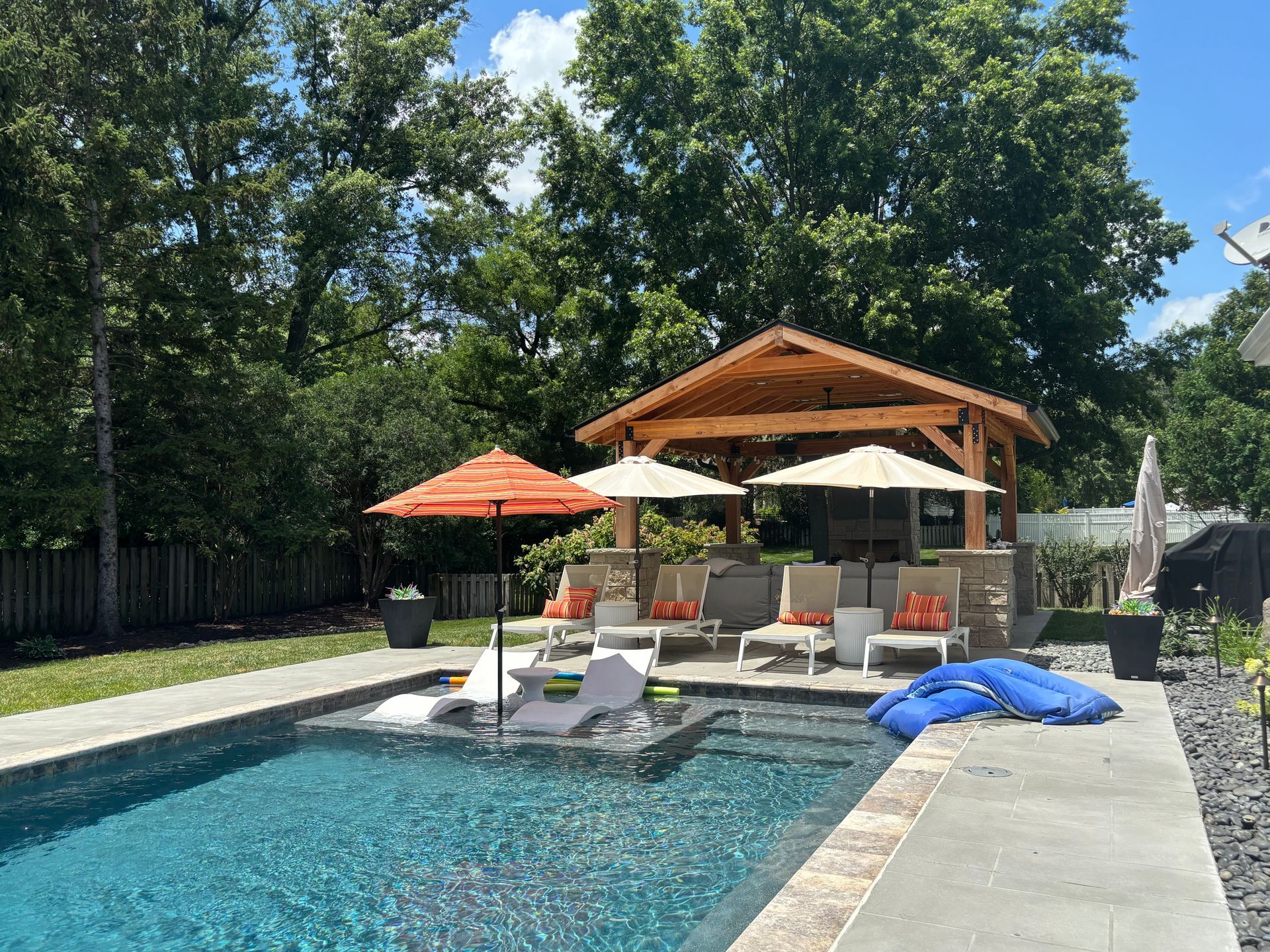Backyard pool with a wooden gazebo, lounge chairs, and umbrellas on a sunny day.