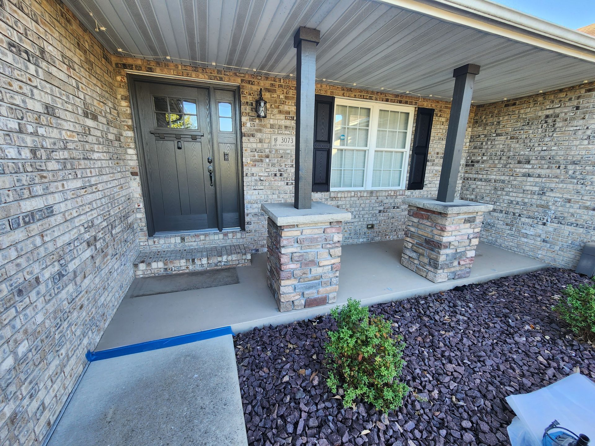 Front porch with stone-covered columns and brick facade. Black door, window with shutters, and small garden.