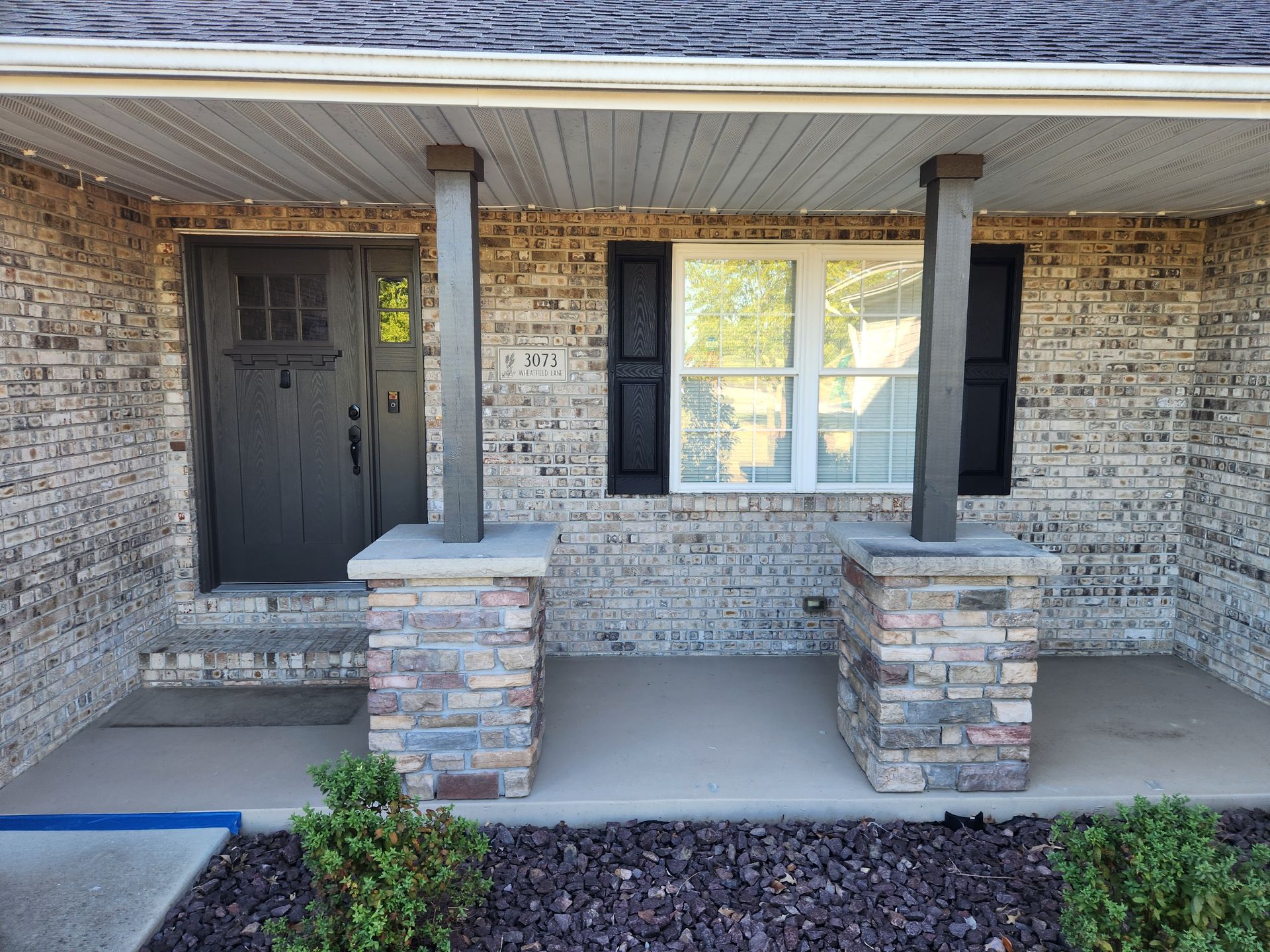 Brick home entryway with stone columns, black door, shutters, and gray porch.