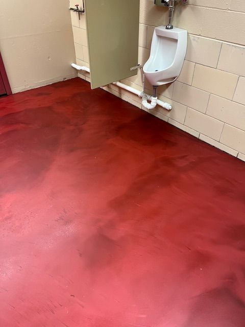 Red floor in a public restroom with a urinal and stall, cream-colored walls, and white plumbing.