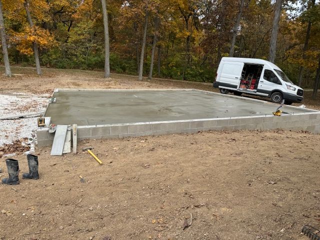 Concrete foundation with cinder block walls, van parked nearby, and tools on site, in a wooded area.