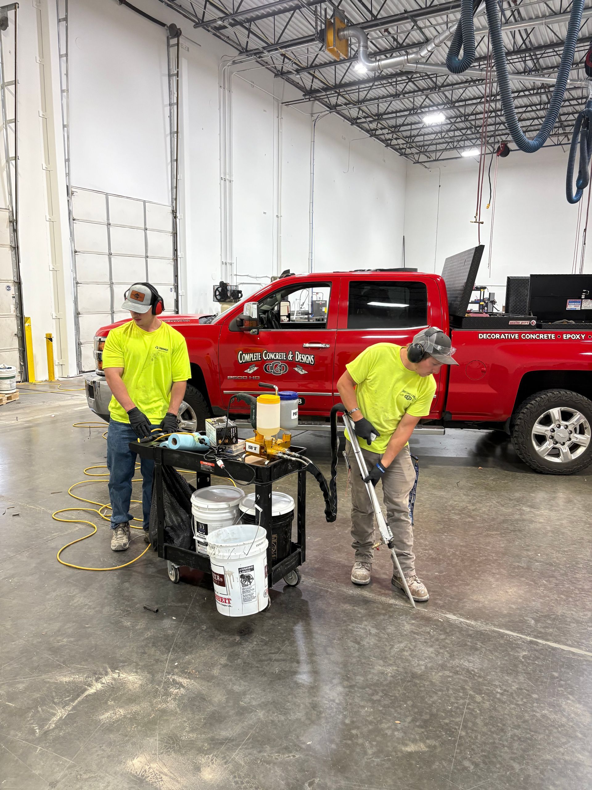 Two workers in neon yellow shirts, one using a jackhammer, another prepping materials near a red truck in a warehouse.