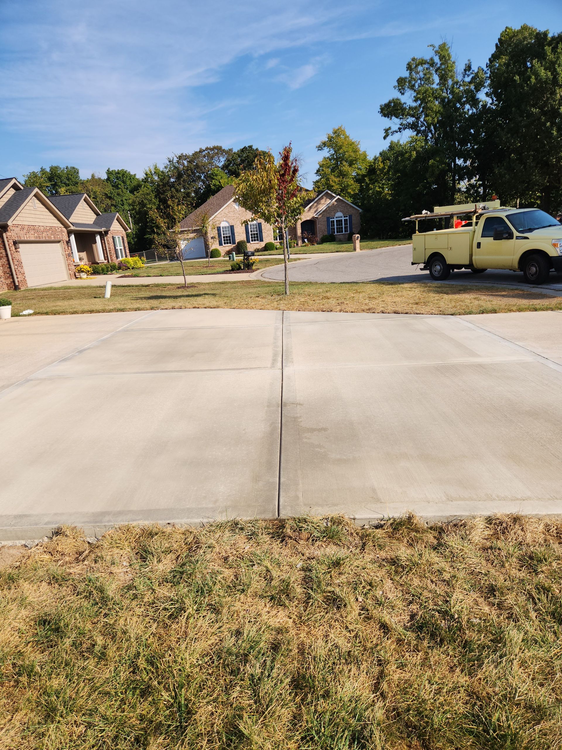 Residential street scene; concrete road with cracked seam, yellow truck, houses, trees, and blue sky.