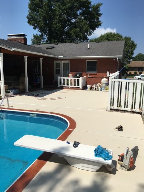Pool area with a diving board, light concrete deck, red brick house, and white fence.
