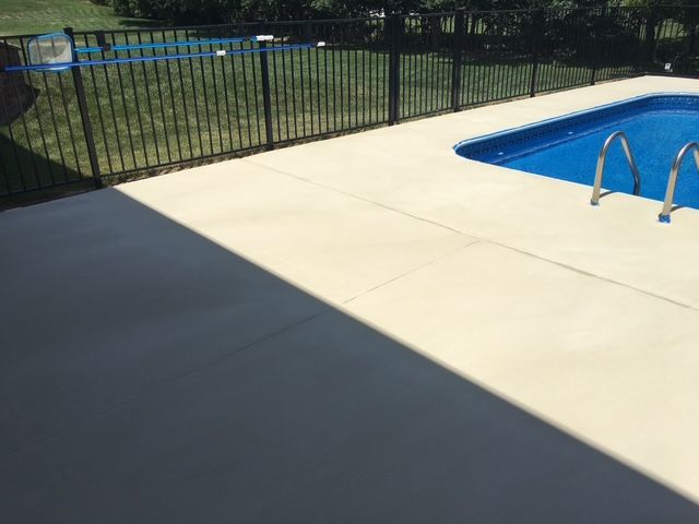Pool deck with cream-colored concrete next to a blue pool, enclosed by a black metal fence and green grass.
