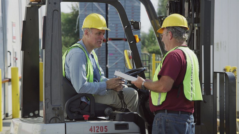 Two construction workers in hard hats exchange paperwork beside a forklift at a worksite.