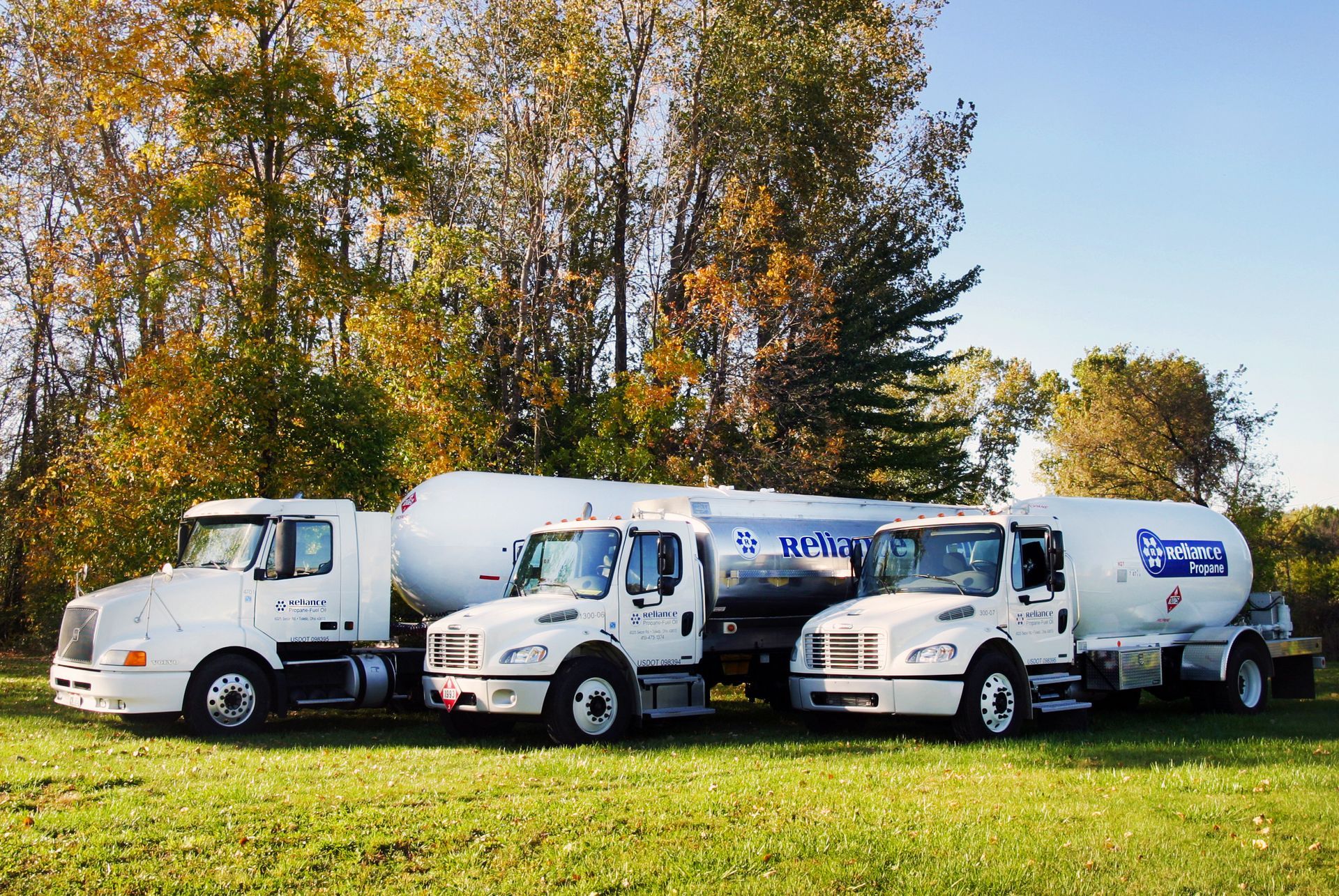 Three white fuel tanker trucks parked on grass with autumn trees in the background