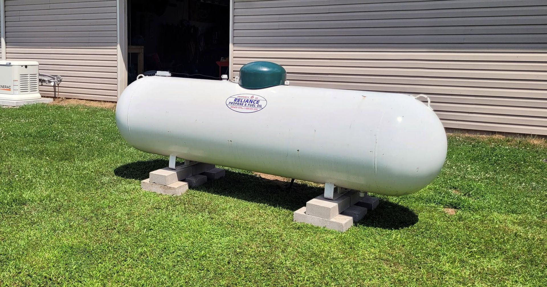 White propane tank on concrete blocks in a grassy yard beside a house.