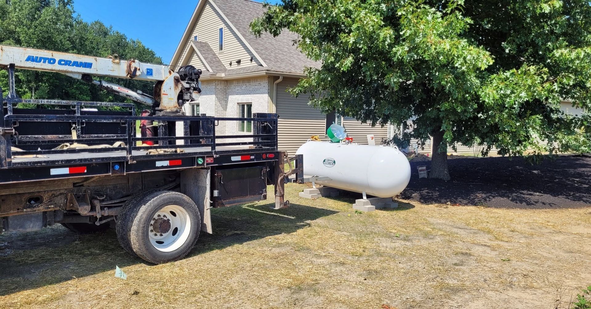 Truck bed beside a house with a white propane tank in a sunny yard.
