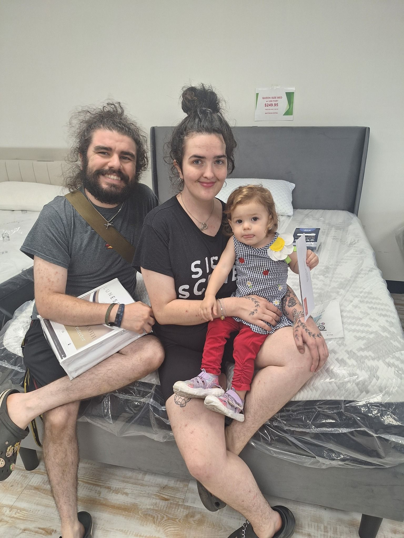 Family of three sits on a bed in a store. Smiling, the woman holds a toddler. Man holds a book.