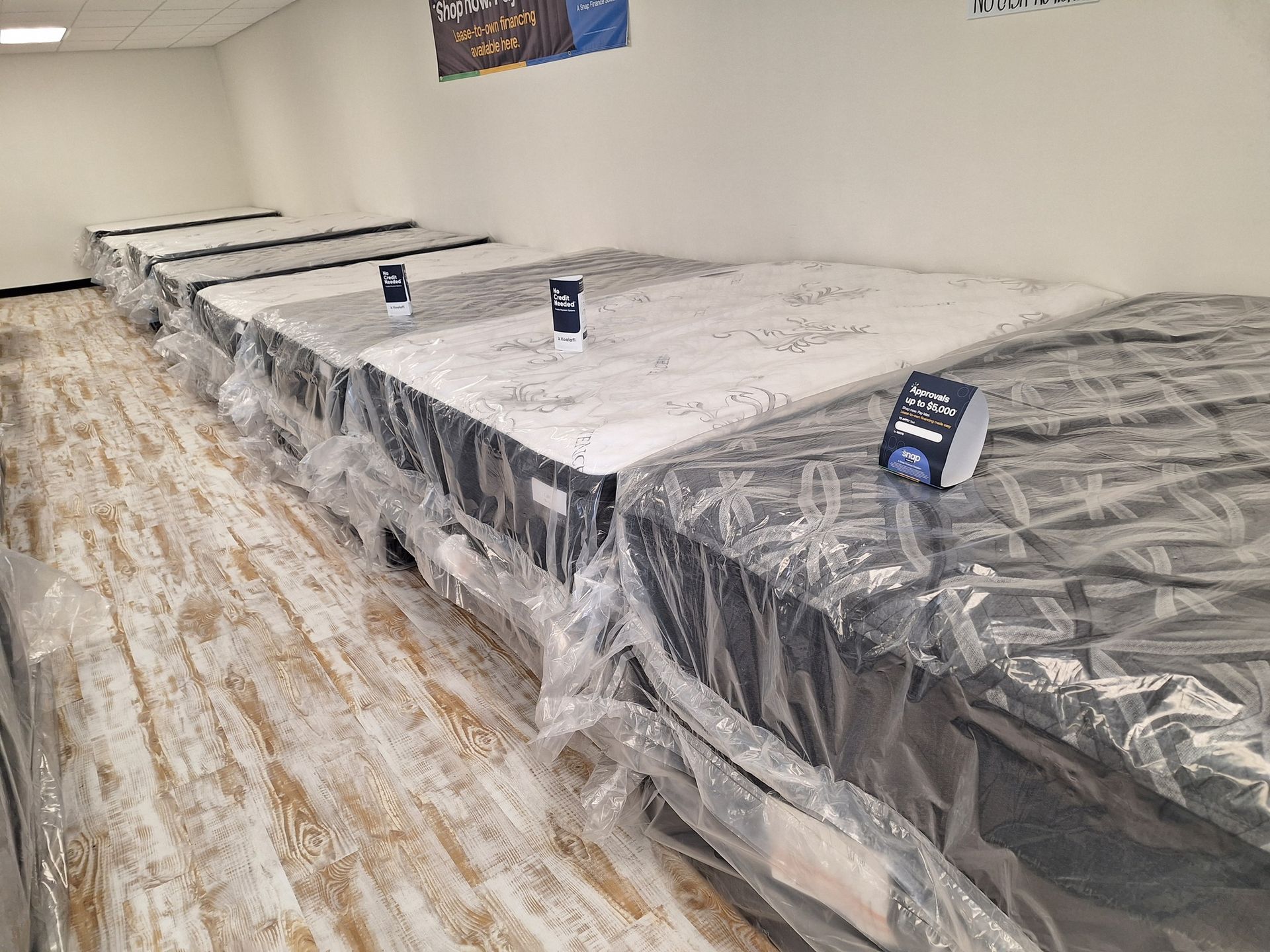Mattresses in plastic wrap displayed in a showroom, set on a light-colored wooden floor.