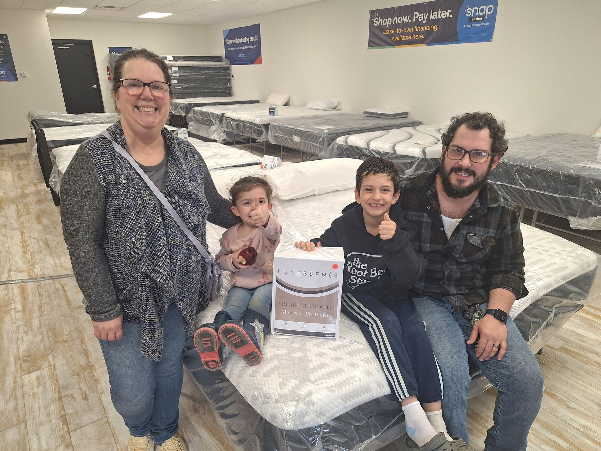 Family in a mattress store, posing with a box; smiling, thumbs up, mattresses in background.