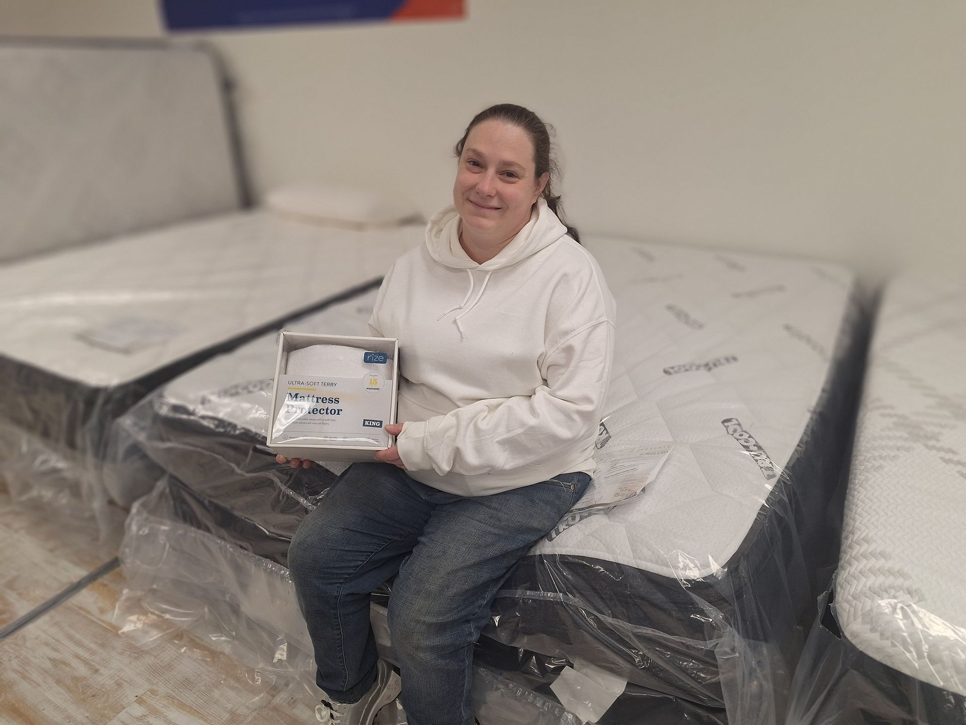 Woman sits on a mattress, holding a product, smiling, in a showroom.