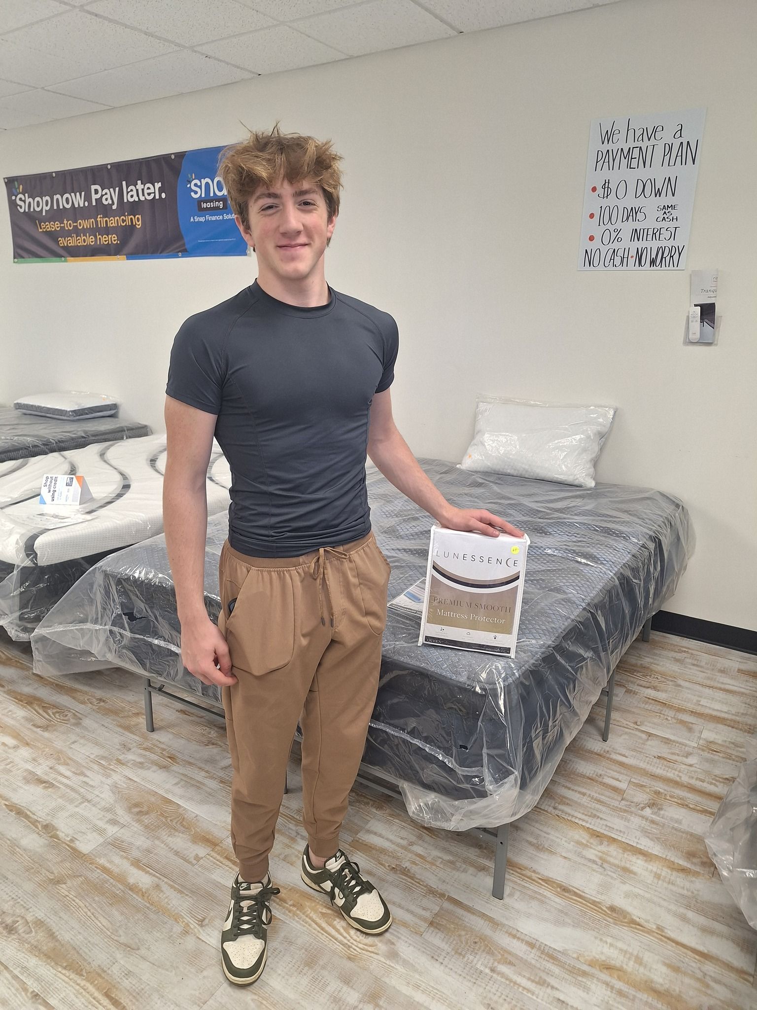 Young man stands near a mattress in a store, holding a box.