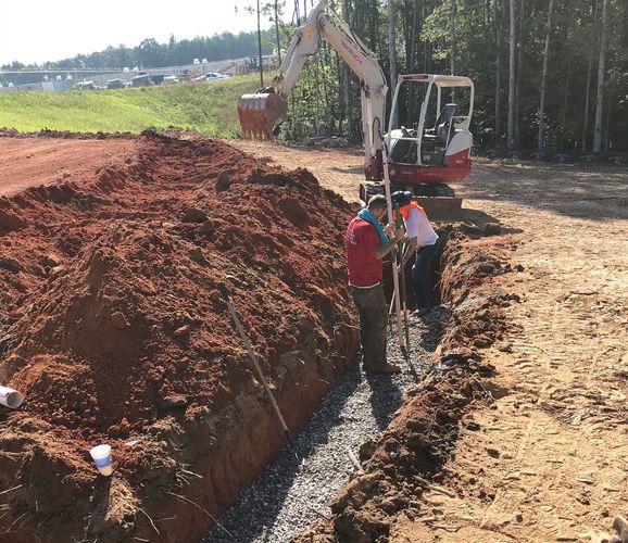 Workers at a dirt excavation site beside a large soil pile and a small excavator.