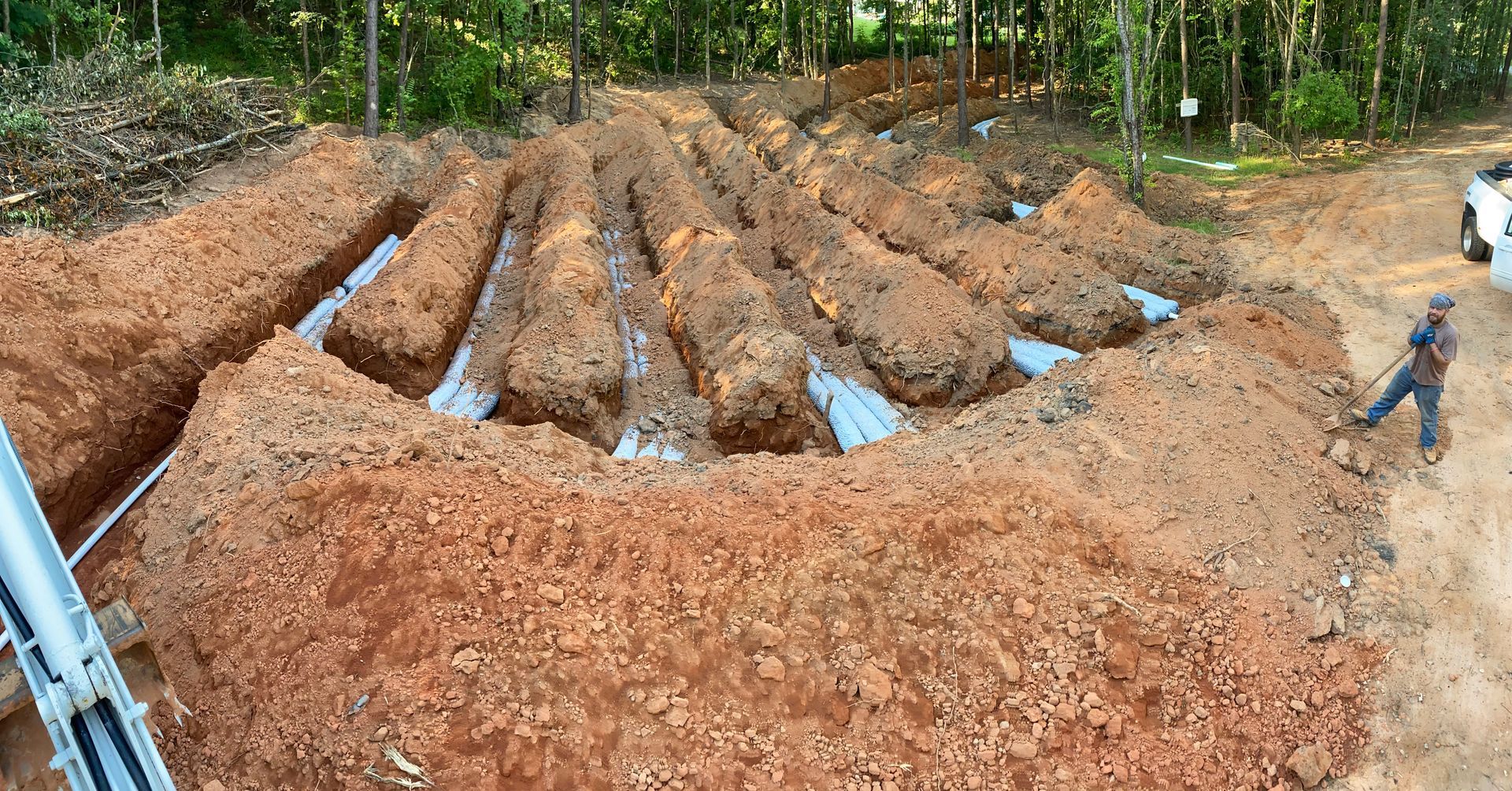 Excavated trenches in a muddy clearing, with workers and a forest edge in the background.