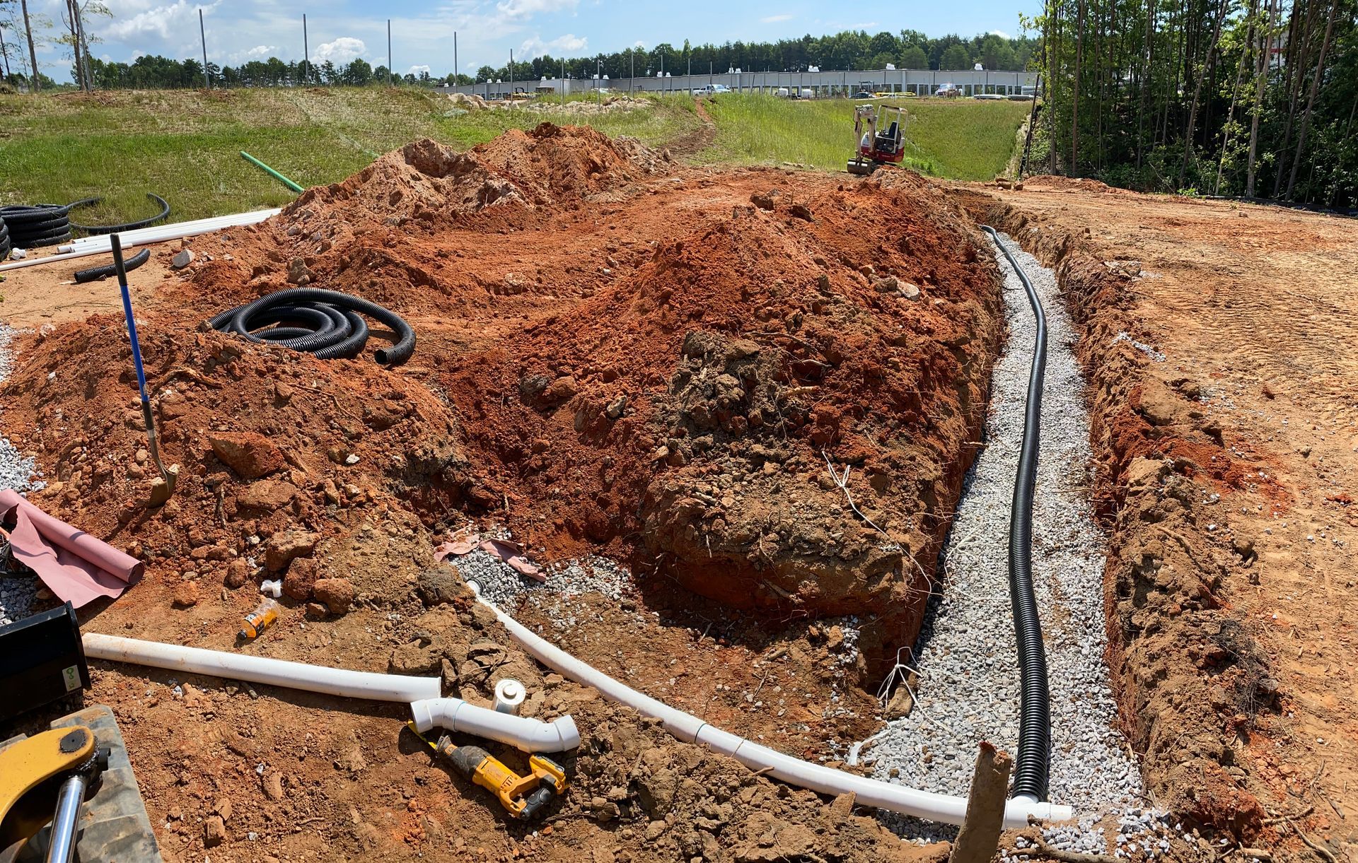 Trenched construction site with exposed black pipe, dirt berms, and a hose under a clear sky.