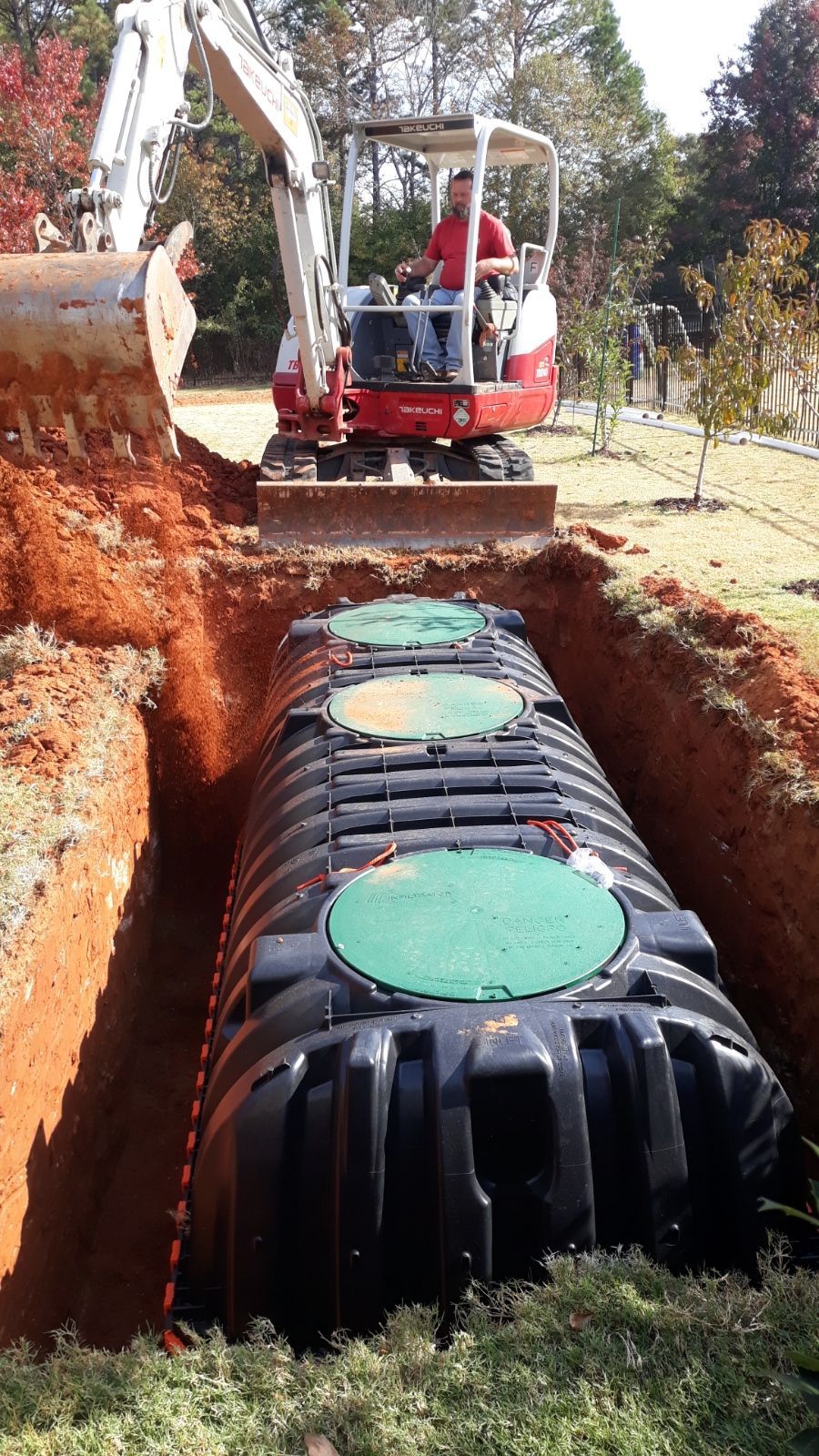 Excavator lowering a large black septic tank into a deep trench on a grassy site.