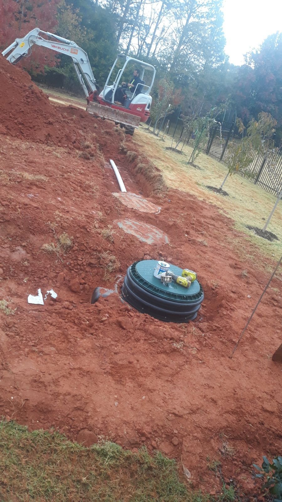 Excavator on a red dirt slope near a black pipe cover in a wooded area.