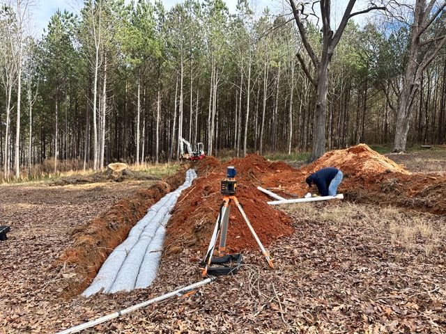 People working on a trench in a wooded area with soil piles and construction materials
