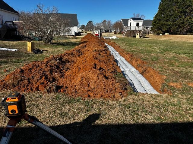 Trenched yard with a long mound of orange dirt and gray drainage pipe along a grassy residential lot.