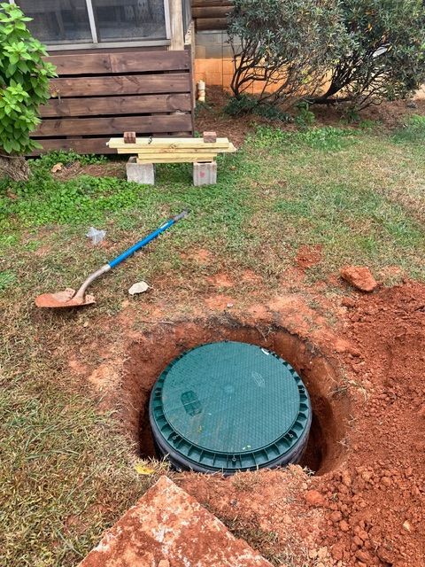 Yard with a dug hole exposing a green septic tank lid beside a house, with a shovel nearby.
