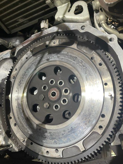 Engine flywheel with visible teeth and bolt holes, likely in an automotive repair setting.
