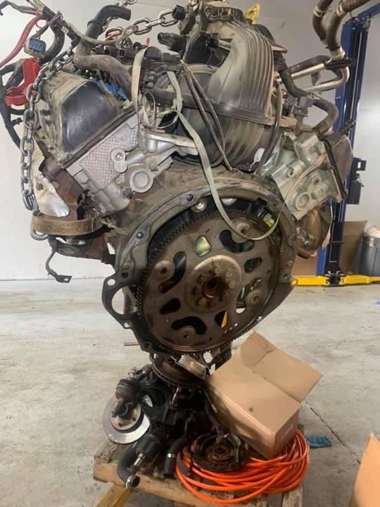 An engine block, heavily assembled, sits on a wooden base with a flywheel exposed. Electrical wires and other parts are around it.