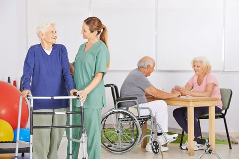 Rehabilitation room with caregivers, an older adult using a walker, and a seated pair beside a wheelchair and exercise balls