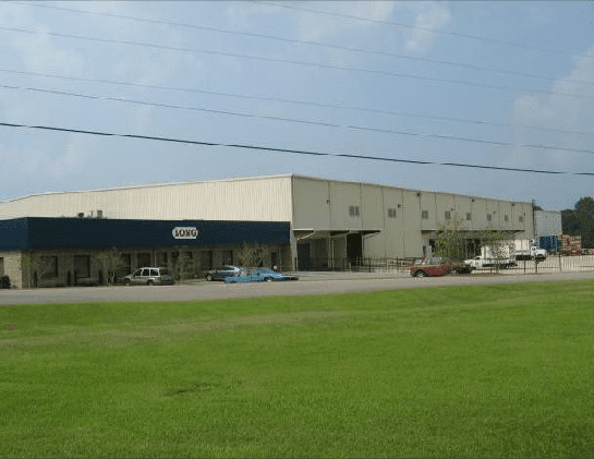 A large white building with a blue roof is surrounded by a lush green field.