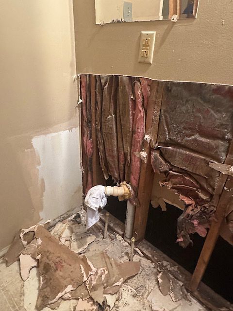 Bathroom wall damaged, revealing insulation and pipes; debris on floor.